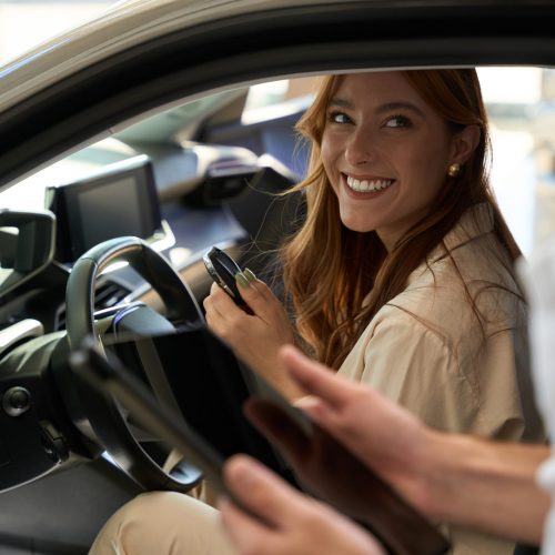 Woman with car key fob sitting at steering wheel and looking at salesclerk with tablet computer in hands